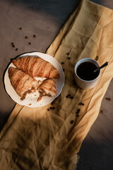 top view white cup of coffee, two croissants on a white plate. broken croissant with chocolate. breakfast on a textured gray surface with a yellow cloth. Morning meal and breakfast concept