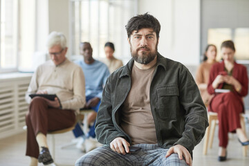 Portrait of serious bearded man dressed in casual wear and looking at camera while posing in office with diverse group of people in background, copy space