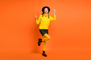 Full size photo of optimistic girl hands fists wear yellow sweater cap skirt shoes isolated on orange background