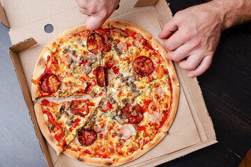 Man hand is cutting an appetizing pizza into slices, in a box, on a black table. Top view.