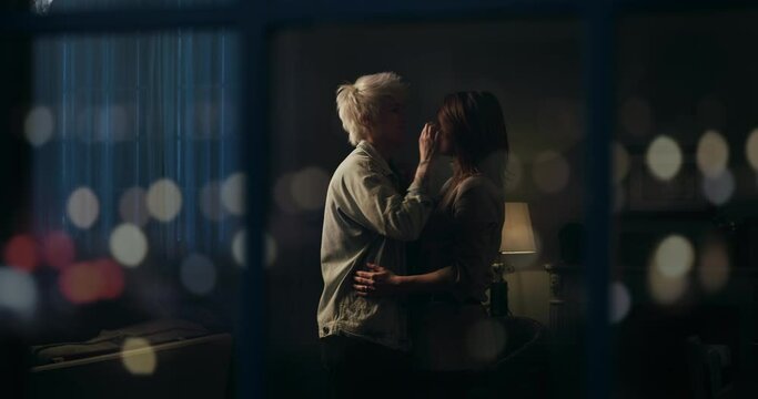 Cinematic shot from window of young happy homosexual female gay and transgender man couple in love enjoying to embrace and kiss while dancing together and listening to the music at home at evening.