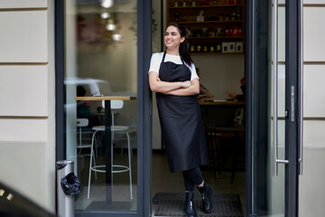 Cheerful brunette caucasian owner of coffee shop in apron standing at entrance greeting visitors...