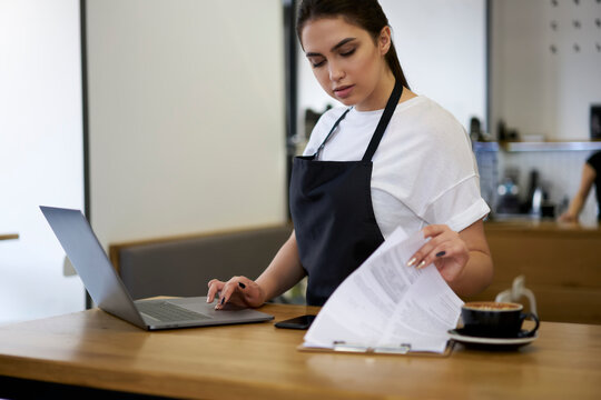 Skilled caucasian woman barista checking invoices and finance of commercial retail in coffee shop during working process, professional female manager planning budget of cafe use laptop computer