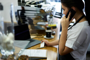 Skilled woman entrepreneur making mobile phone planning business service support at working place with technology, young female barista holding visit card booking equipment via call for coffee shop