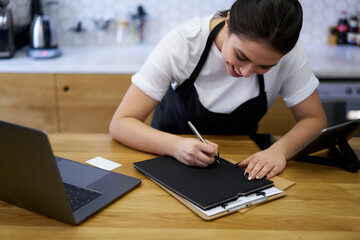 Cropped image of skilled concentrated female manager of coffee shop writing finance report and income from business near laptop computer, skilled woman barista filling