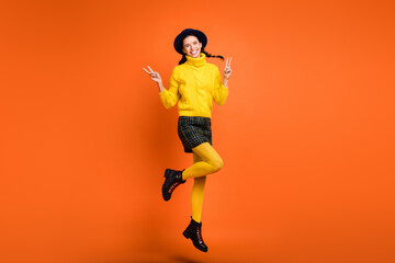 Full length body size view of pretty cheerful girl jumping showing v-sign having fun isolated over bright orange color background