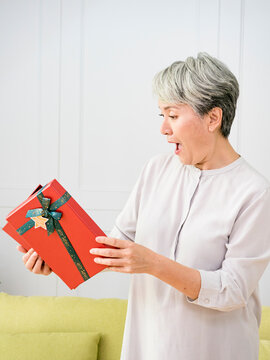 Portrait Of An Elderly Asian Woman Receives Gift Boxes, Smiling And Looking Down While Standing In A Living Room At Home.