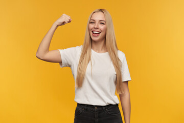 Teenage girl, happy looking woman with blond long hair. Wearing white t-shirt and black jeans. Shows her muscles Watching at the camera, isolated over orange background