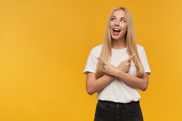 Nice looking woman, beautiful girl with long blond hair. Wearing white t-shirt and black jeans. Watching to the left and pointing both sides at copy space, isolated over orange background