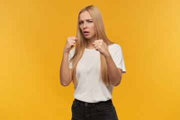 ready to fight, happy looking woman with blond long hair. Wearing white t-shirt and black jeans. People and emotion concept. Watching at the camera, isolated over orange background
