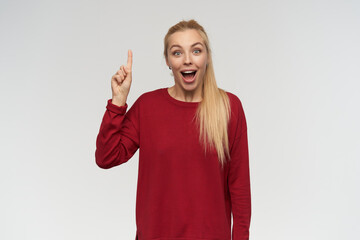 Portrait of attractive, adult girl with blond long hair. Wearing red sweater. People and emotion concept. Watching at the camera, keeps her finger up isolated over white background