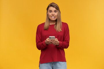 Blonde young woman looks insecure as if she's in trouble or an awkward situation, keeps mobile phone in her hands, looking camera with fear, bites slightly her lip, on a yellow studio background