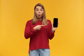 Blonde girl looks amazed, mouth rounded in excitement, keeps mobile phone in her hand, black screen turned to the camera, points on it with index finger,standing isolated on a yellow studio background