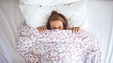 Funny young woman lying in bed and hiding under sheet while looking up with copy space. Top view of girl hiding face under white blanket on bed in the morning. 