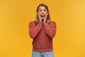 Blonde young surprised and shocked with great news, keeps palms near cheeks, standing isolated on a...