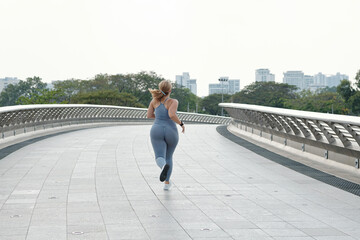 Plus size young woman jogging on empty bridge early in the morning, view from the back, determination concept