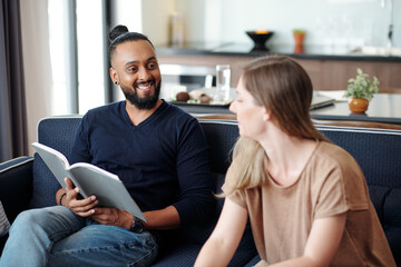 Smiling handsome mixed-race man with open book looking at talking wife