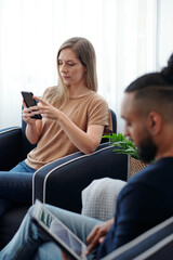 Serious pretty young woman sitting in armchair next to husband and answering text messages on smartphone or reading article