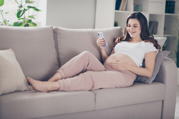 Full size photo of young beautiful lovely happy positive smiling pregnant woman lying on coach browsing in smartphone at home