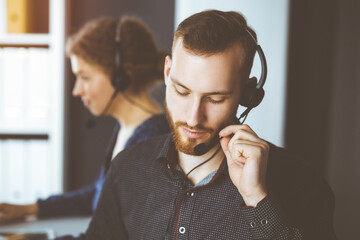 Red-bearded businessman talking by headset near his female colleague while sitting in sunny office. Diverse people group in call center