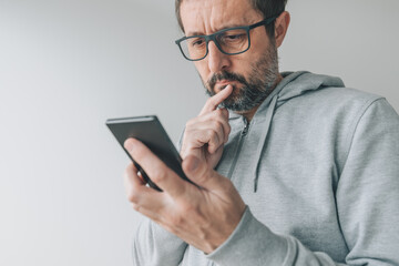 Serious thoughtful mid-adult male looking at mobile smart phone screen with finger on lips