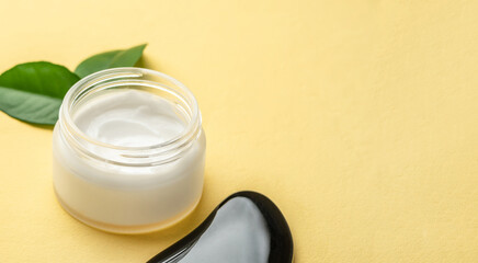 Container of cream and green leaves on a yellow table with copy space. Concept of facial skin care and the use of cosmetics with a natural organic composition