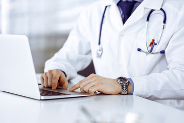 Unknown male doctor sitting and working with laptop in clinic at his working place, close-up. Young physician at work. Perfect medical service, medicine concept