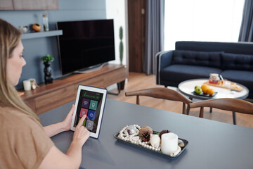 Woman sitting at table in studio apartment and usig smart home application on digital tablet