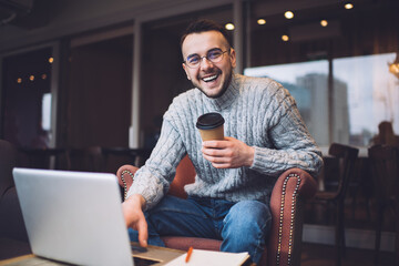 Cheerful man with laptop and cup of coffee in armchair