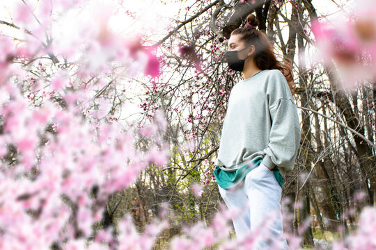 Portrait of young woman in protective face mask standing under cherry blossoms