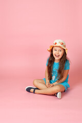 full-length portrait of a crying, horrified, open-mouthed girl with long wavy hair in a straw hat and blue dress sitting cross-legged on the floor, isolated on a pink background.