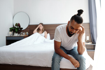 Unhappy bearded man sitting on bed edge after having argument with wife