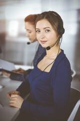 Two busineswomen have conversations with the clients by headsets, while sitting at the desk in a...