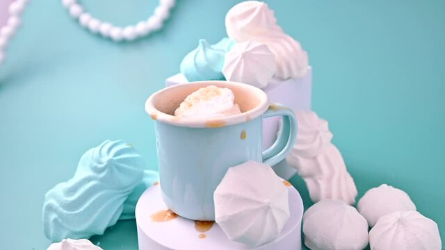 A meringue figurine falls into a cup of black coffee and splashes fly. Espresso with froth and meringues of different colors on a blue background. A creative shot of protein and sugar cookies. 