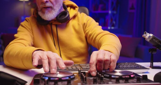 Close up of cool stylish Caucasian bearded senior man DJ sitting at desk in dark room with neon lights working on console mixer equipment mixing new dance music, sound beat maker