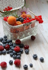 colorful forest fruits, small apricots and flowers, arranged raspberries, red currants and blueberries, natural vitamins, glass box filled with fruit, old, retro box made of hewn glass