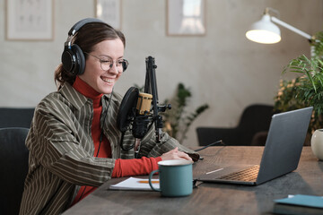 Young reporter in headphones sitting at the table looking at monitor of laptop and talking online in the studio