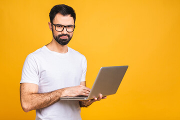 Confident business expert. Confident young handsome bearded man in casual holding laptop and smiling while standing over isolated yellow background.