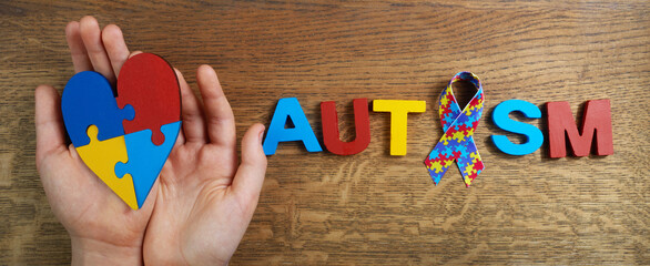 Autistic boy hands holding heart shapeed puzzle with word autism and awarennes ribbon on wooden background. Autism awareness day or month.