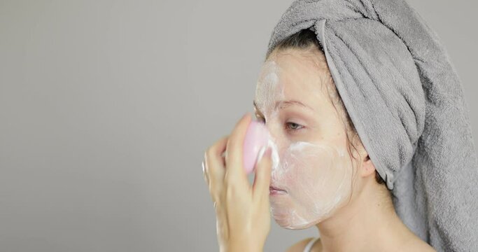 Side view of beautiful charming young girl with towel on her head washing off cosmetic mask from her face using silicone brush while looking in the mirror. Home skin care concept