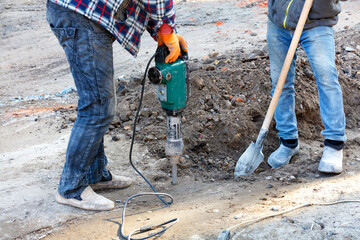 Builders at a construction site are loosening the dense soil with an electric jackhammer and shovel.