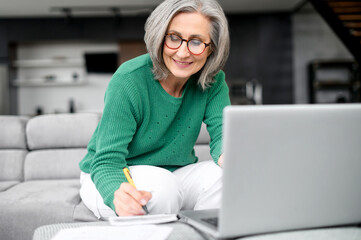 Confident mature woman using laptop computer for remote work, watching webinar and taking notes sitting at home. Contemporary senior female online teacher holding video conference and making marks - obrazy, fototapety, plakaty