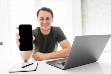 Happy handsome man freelancer sitting at the kitchen table, working on laptop computer, showing blank screen phone