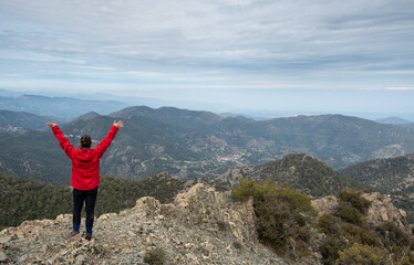 Tourist woman with raised arms standing on rocky top against a cloudy sky enjoying mountain range panorama.