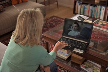 A woman sits in front of her laptop at home, talking with colleagues on a video call and watching...