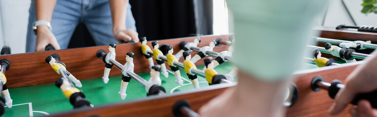 partial view of interracial business colleagues playing table football, blurred foreground, banner