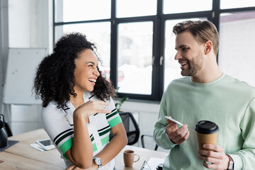 Fototapeta premium happy businessman holding coffee and smartphone while talking to laughing african american colleague