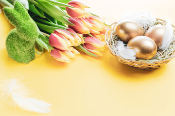 Basket easter decoration: Golden eggs in basket with spring tulips, white feathers on pastel yellow background. Congratulatory easter design.
