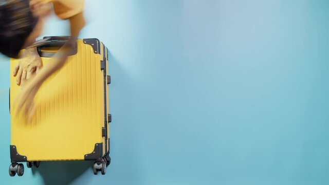 Timelapse – Asian black hair woman packs bright yellow suitcase with face mask and alcohol sanitizer to protect covid-19 for optimistic summer holidays on blue background. Top view with copy space.