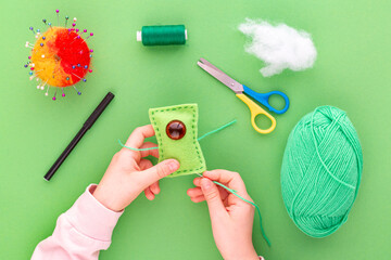 Instruction step by step, step nine. The hands of a child in the process of sewing a toy from felt, a green one-eyed monster. Green background, flat lay. Hobbies, handicrafts, fine motor skills.